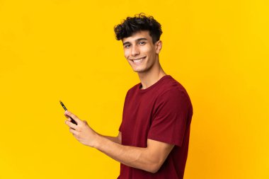 Arjantinli genç adam sarı arka planda izole edilmiş cep telefonuyla mesaj ya da e-posta gönderiyor.