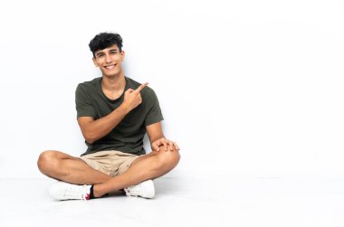 Yerde oturan genç Arjantinli bir adam bir ürünü sunmak için kenarı işaret ediyor.