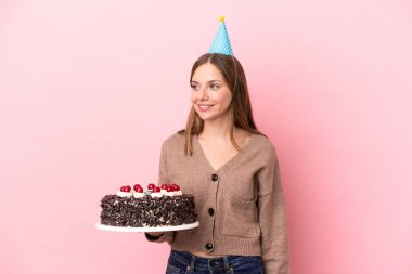 Elinde doğum günü pastası tutan Litvanyalı genç bir kadın pembe arka planda izole edilmiş bir şekilde yan tarafa bakıyor ve gülümsüyor.