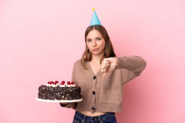 Elinde doğum günü pastası tutan Litvanyalı genç bir kadın pembe arka planda izole edilmiş, olumsuz bir ifadeyle baş parmağını gösteriyor.