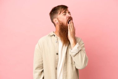 Kırmızımsı beyaz adam pembe arka planda izole edilmiş esniyor ve geniş ağzını eliyle kapatıyor.