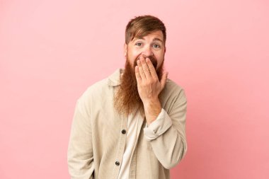 Kırmızımsı beyaz adam pembe arka planda izole edilmiş mutlu ve gülümseyen ağızları eliyle örtüyor.