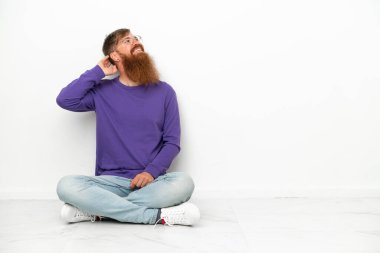 Beyaz arka planda izole bir şekilde yerde oturan Kafkasyalı genç bir adam bir fikir düşünüyor.