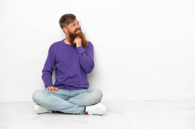 Beyaz arka planda izole bir şekilde yerde oturan Kafkasyalı genç adam yan tarafa bakıyor.