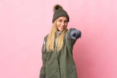 Kış şapkalı genç kadın pembe arka planda izole edilmiş mutlu bir ifadeyle ön tarafı işaret ediyor.