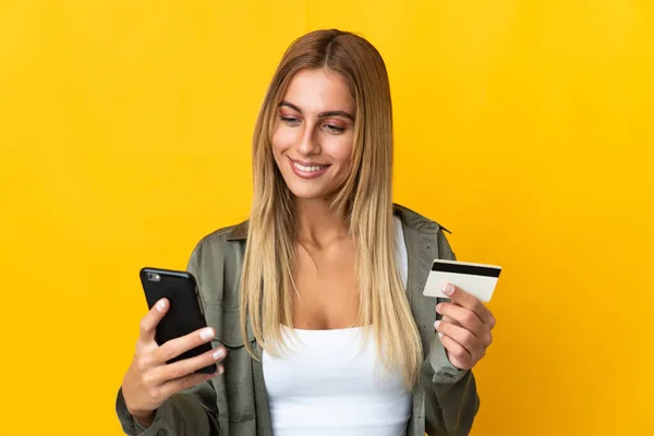 Młoda Blondynka Odizolowana Żółtym Tle Kupuje Komórką Kartą Kredytową — Zdjęcie stockowe
