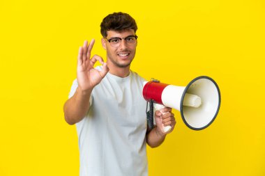 Sarı arka planda izole edilmiş beyaz, yakışıklı genç bir adam elinde bir megafonla tamam işaretini parmaklarıyla gösteriyor.