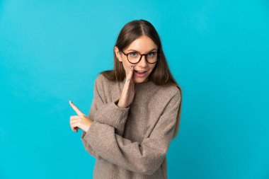 Mavi arka planda izole edilmiş genç Litvanyalı kadın bir ürünü sunmak ve bir şeyler fısıldamak için kenarı işaret ediyor.