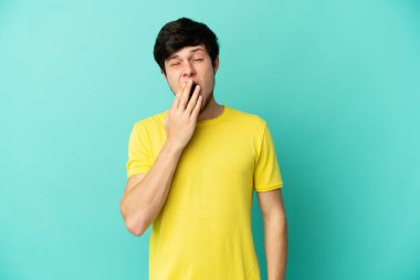 Mavi arka planda izole edilmiş genç bir Rus adam esniyor ve elleriyle ağzını kapıyor.