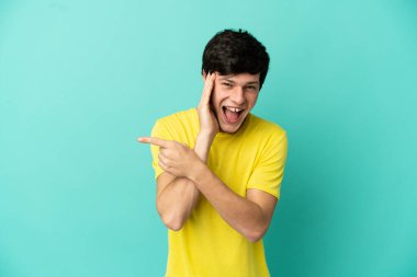 Mavi arka planda izole edilmiş genç bir Rus bir ürünü sunmak ve bir şeyler fısıldamak için kenarı işaret ediyor.