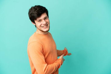 Genç Rus adam mavi arka planda izole edilmiş. Davet etmek için ellerini yana uzatıyor.
