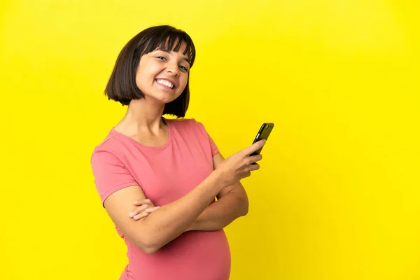 Młoda Mieszana Rasa Ciężarna Kobieta Odizolowana Żółtym Tle Trzymając Telefon — Zdjęcie stockowe