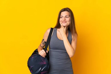 Sarı arka planda izole edilmiş spor çantalı Slovak kadın yana bakıyor ve gülümsüyor.