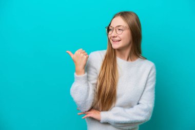 Mavi arka planda izole edilmiş genç bir Rus kadın bir ürünü sunmak için kenarı işaret ediyor.