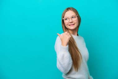 Mavi arka planda izole edilmiş genç bir Rus kadın bir ürünü sunmak için kenarı işaret ediyor.