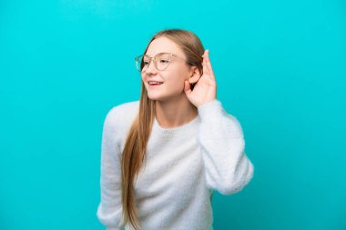 Genç Rus kadın mavi arka planda izole edilmiş. Kulağına elini sürerek bir şeyler dinliyor.