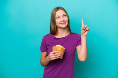 Mavi arka planda izole edilmiş patates kızartması yiyen genç Rus kadın harika bir fikre işaret ediyor.