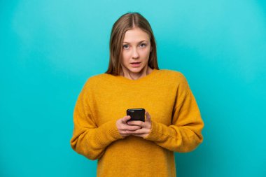 Genç Rus kadın mavi arka planda izole edilmiş kameraya bakarken şaşırmış bir ifadeyle cep telefonunu kullanıyor.