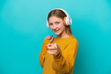 Genç Rus kadın mavi arka planda izole edilmiş müzik dinliyor ve ön tarafı işaret ediyor.
