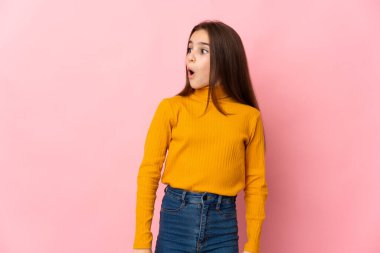 Pembe arka planda izole edilmiş küçük bir kız yan tarafa bakarken sürpriz jest yapıyor.