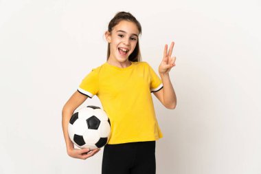 Beyaz arka planda izole edilmiş küçük futbolcu kız gülümsüyor ve zafer işareti gösteriyor.