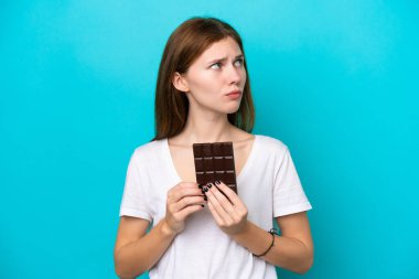Genç İngiliz kadın mavi arka planda izole edilmiş çikolata tableti alıyor ve kuşkuları var.