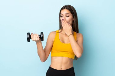 Beyaz kadın halter yapıyor. Mavi arka planda izole edilmiş. Şaşırmış ve şok olmuş bir yüz ifadesi var.