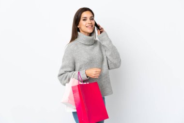 Beyaz arka planda izole edilmiş genç beyaz bir kadın alışveriş torbaları tutuyor ve cep telefonuyla arkadaşını arıyor.
