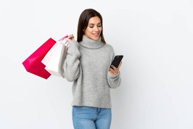 Beyaz arka planda izole edilmiş genç beyaz bir kadın alışveriş torbaları tutuyor ve bir arkadaşına cep telefonuyla mesaj yazıyor.
