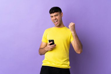 Mor arka planda izole edilmiş genç Kolombiyalı adam telefon zafer pozisyonunda.