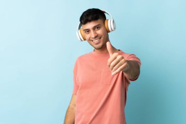 Genç Kolombiyalı adam mavi arka planda izole edilmiş müzik dinliyor ve başparmağını kaldırıyor.