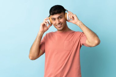 Genç Kolombiyalı adam mavi arka planda izole edilmiş müzik dinliyor.
