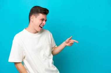 Mavi arka planda izole edilmiş genç ve yakışıklı Brezilyalı bir adam parmak göstererek bir ürün sunuyor.