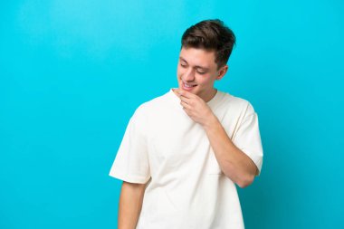 Genç ve yakışıklı Brezilyalı bir adam mavi arka planda izole edilmiş, yana bakıyor ve gülümsüyor.