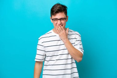 Genç, yakışıklı Brezilyalı bir adam mavi arka planda izole edilmiş mutlu ve gülümseyen bir ağızla elini kapatıyor.