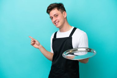 Tepsisi mavi arka planda izole edilmiş genç bir garson parmak kenarı işaret ediyor.