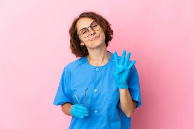 İngiliz dişçi kadın, pembe arka planda izole edilmiş aletleri tutuyor mutlu ve parmaklarıyla dört tane sayıyor.