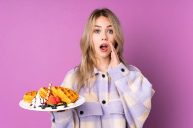 Elinde waffle tutan genç bir Rus kız. Mor arka planda izole edilmiş. Şaşırmış ve şok olmuş bir yüz ifadesi var.