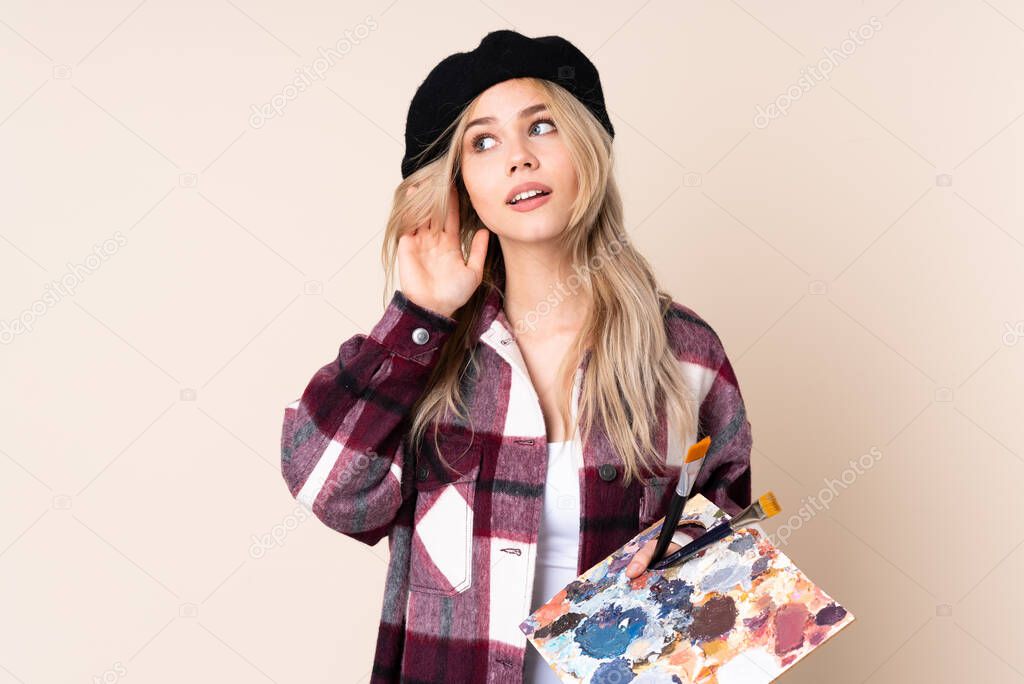 Teenager artist girl holding a palette isolated on blue background listening to something by putting hand on the ear