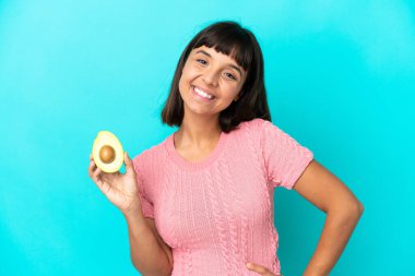 Young mixed race woman holding an avocado isolated on blue background posing with arms at hip and smiling