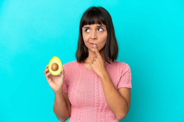 Young mixed race woman holding an avocado isolated on blue background having doubts while looking up