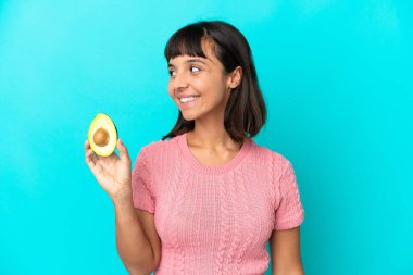 Young mixed race woman holding an avocado isolated on blue background looking side