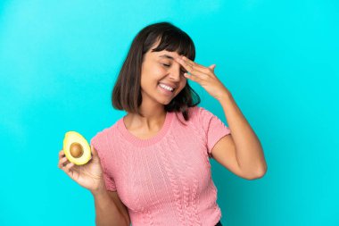 Young mixed race woman holding an avocado isolated on blue background smiling a lot