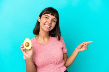 Young mixed race woman holding an avocado isolated on blue background pointing finger to the side