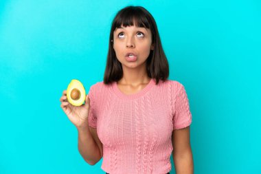 Young mixed race woman holding an avocado isolated on blue background looking up and with surprised expression