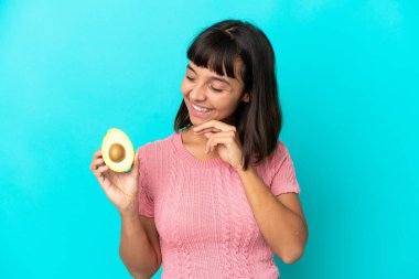 Young mixed race woman holding an avocado isolated on blue background looking to the side and smiling