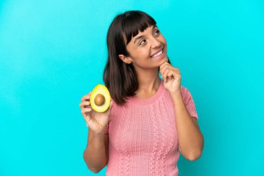 Young mixed race woman holding an avocado isolated on blue background and looking up