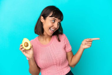 Young mixed race woman holding an avocado isolated on blue background pointing finger to the side and presenting a product