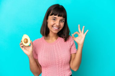 Young mixed race woman holding an avocado isolated on blue background showing ok sign with fingers