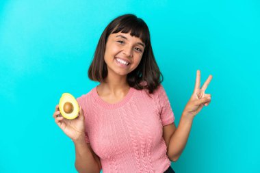 Young mixed race woman holding an avocado isolated on blue background smiling and showing victory sign